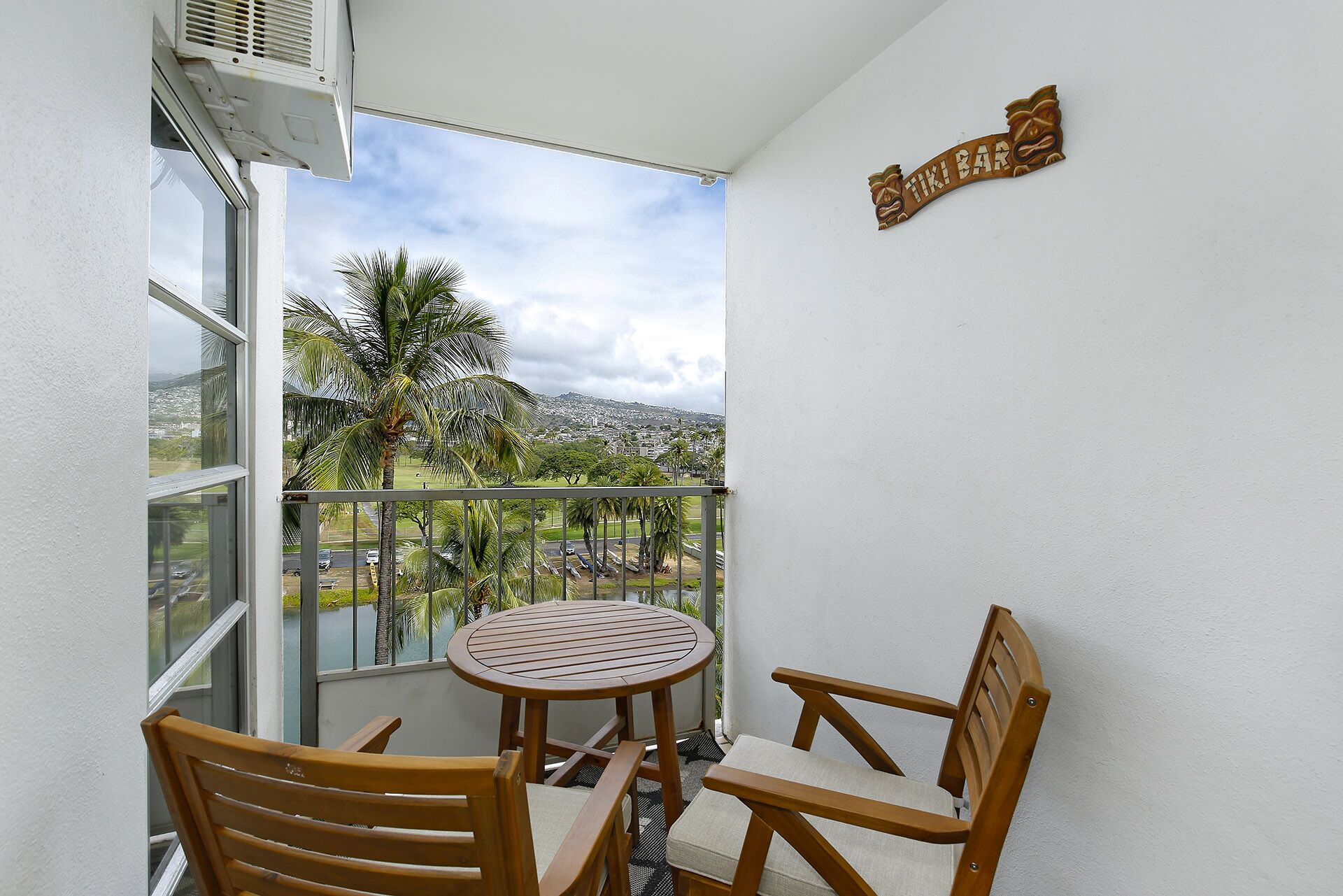 Een balkon met een houten tafel en stoelen, uitzicht op een stadsbeeld, en een bord waarop staat 