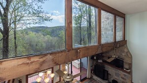 A room with large windows overlooking a forested area, a stone fireplace, and a ceiling fan. - Eagle Point*VIEWS*Fireplace*Sleeps4*Resort*HIke* (Deep Gap)