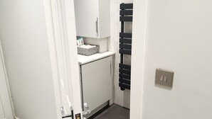 Kapea käytävä, jossa on valkoinen ovi, pieni keittiötila, jossa on työtaso ja kaappeja, sekä musta pyyhekoukku.