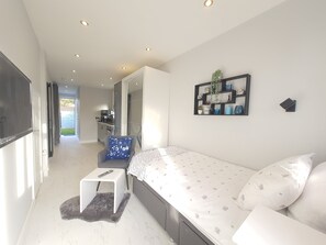 Ein modernes Schlafzimmer mit einem Bett, einer kleinen Sitzecke, einem Fernseher und einer Küche im Hintergrund.