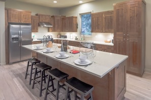A modern kitchen with wooden cabinets, a stainless steel refrigerator, a central island with bar stools, and a window with a view of trees. - Private Hot Tub ~ Luxury Home ~ Close to Bachelor~ Golf (Bend)