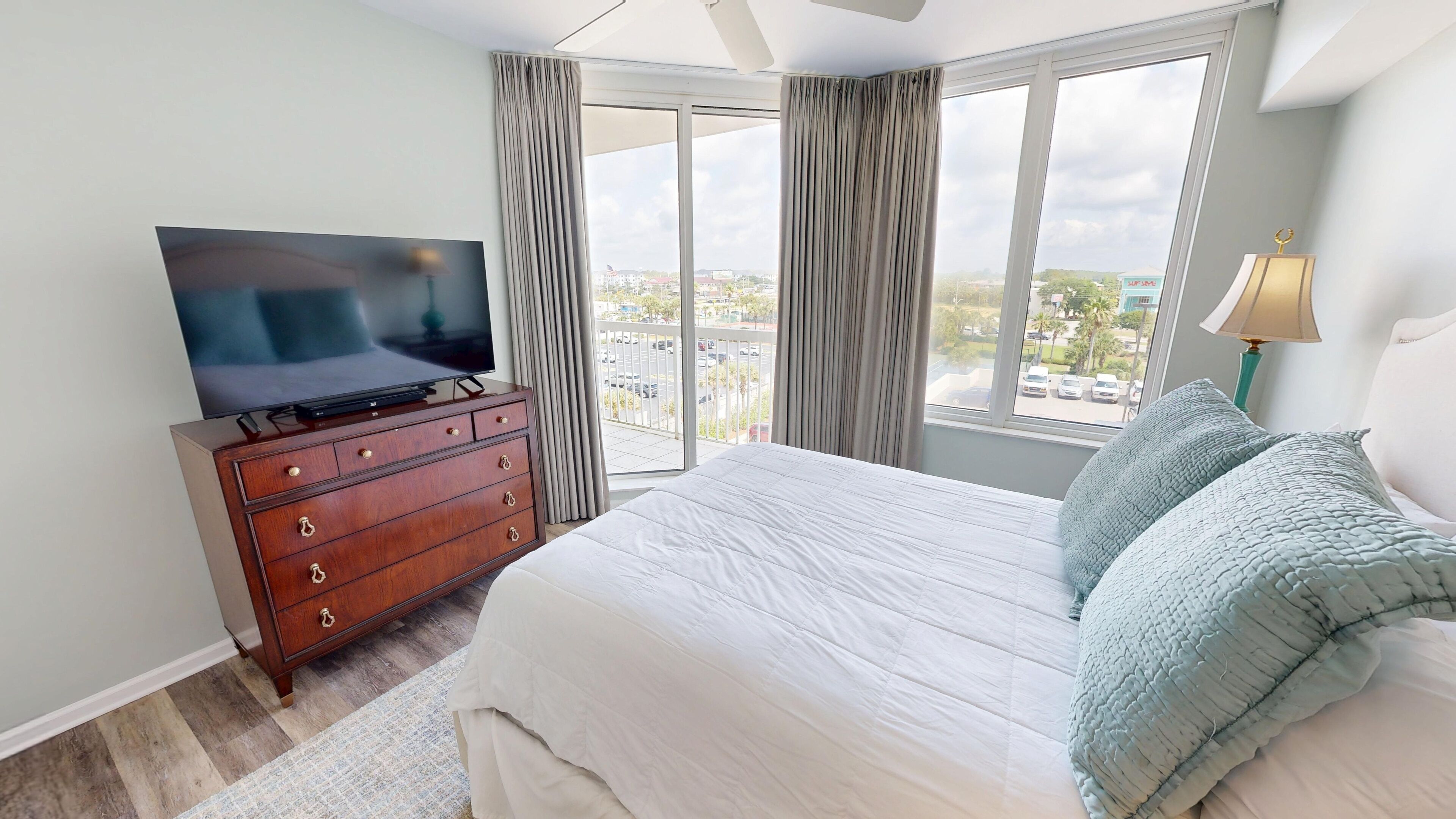 A hotel room with a large bed, a TV on a wooden dresser, a balcony with curtains, and a view of the city.