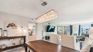 A modern living room with a wooden dining table, comfortable seating, a large TV, and a view of the outside. - IR 518 is a Stunning 2 BR that has amazing Sunset views - sleeps 10 by RedAwning (Destin)