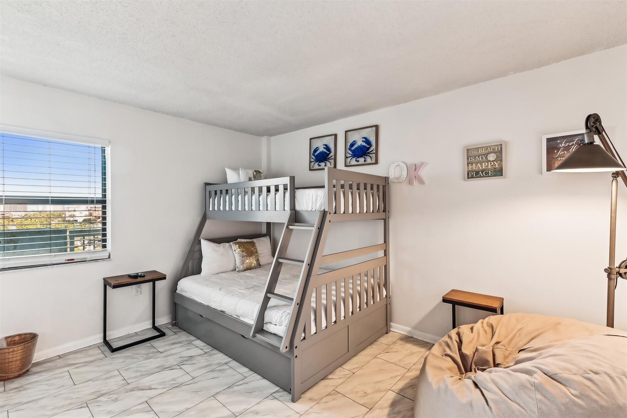 A bedroom with a bunk bed, a desk, a lamp, and a view of the outside.