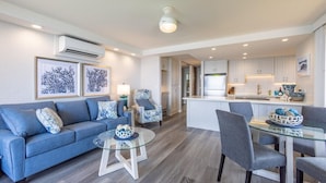 A modern living room with a blue sofa, a glass coffee table, and a dining area with grey chairs. - Papakea L-301  Direct Oceanfront Corner Unit with Panoramic Views (Lahaina)