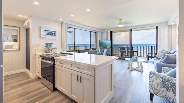A modern kitchen with a large island, a dining area with a view, and a balcony with a railing. - Papakea L-301  Direct Oceanfront Corner Unit with Panoramic Views (Lahaina)