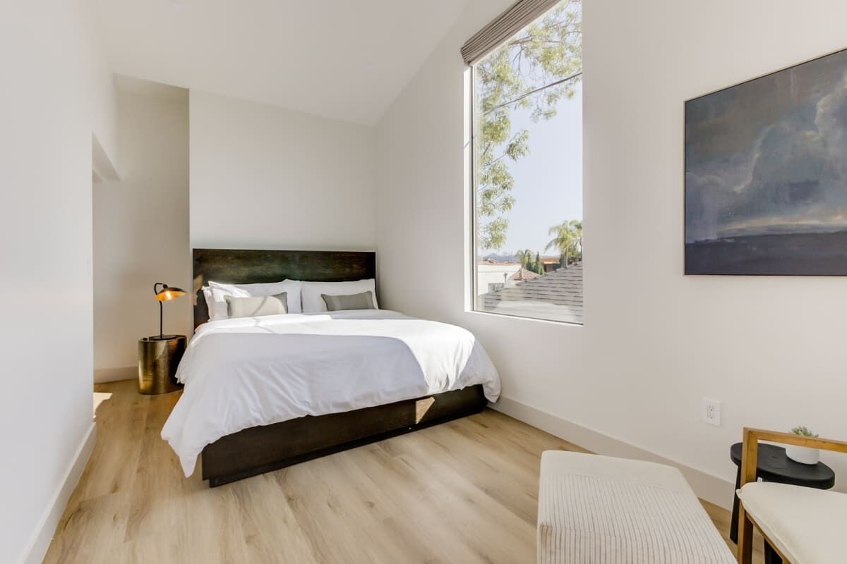 A modern bedroom with a large bed, a bedside table with a lamp, a bench, and a view of the outdoors through a window.