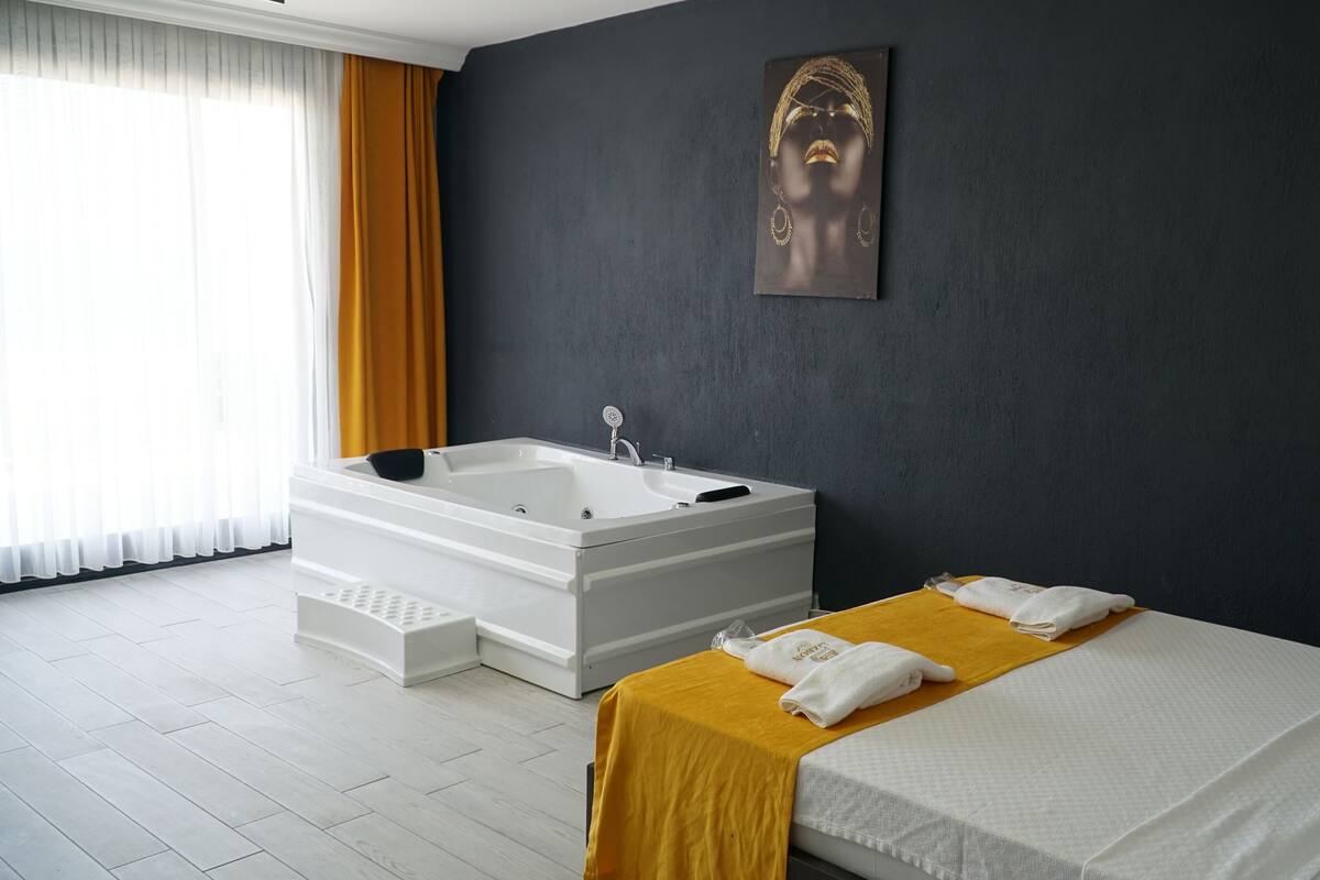A modern hotel room with a large white bathtub, a bed with a yellow towel, and a dark wall with a framed picture.