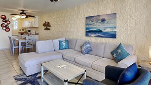 A living room with a sectional sofa, a coffee table, and a blue armchair. There is a dining area with a table and chairs, and a kitchen in the background. - Where Blue Waters Beckon Maalaea Remodeled 2BR Condo Ocean Views Harbor (Maalaea)