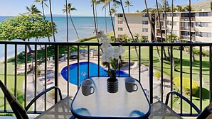 A balcony with a table, two chairs, and a vase with white flowers. The view includes a pool, palm trees, and a building with multiple floors. - Where Blue Waters Beckon Maalaea Remodeled 2BR Condo Ocean Views Harbor (Maalaea)
