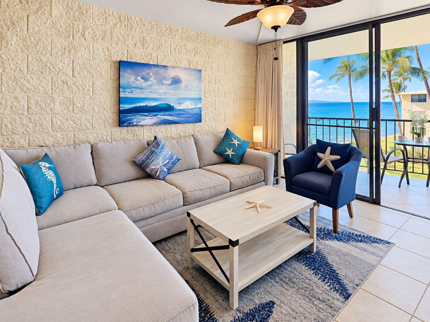 A living room with a sofa, armchair, and coffee table, a painting of a beach scene, and a view of the ocean through large windows.