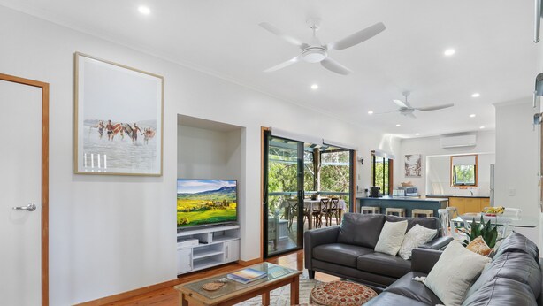 A modern living room with a leather sofa, a coffee table, and a flat-screen TV. There are ceiling fans, a kitchen area, and a dining space visible. - Large, pets welcome family home close to beaches, shops and public transport. (Point Lookout)