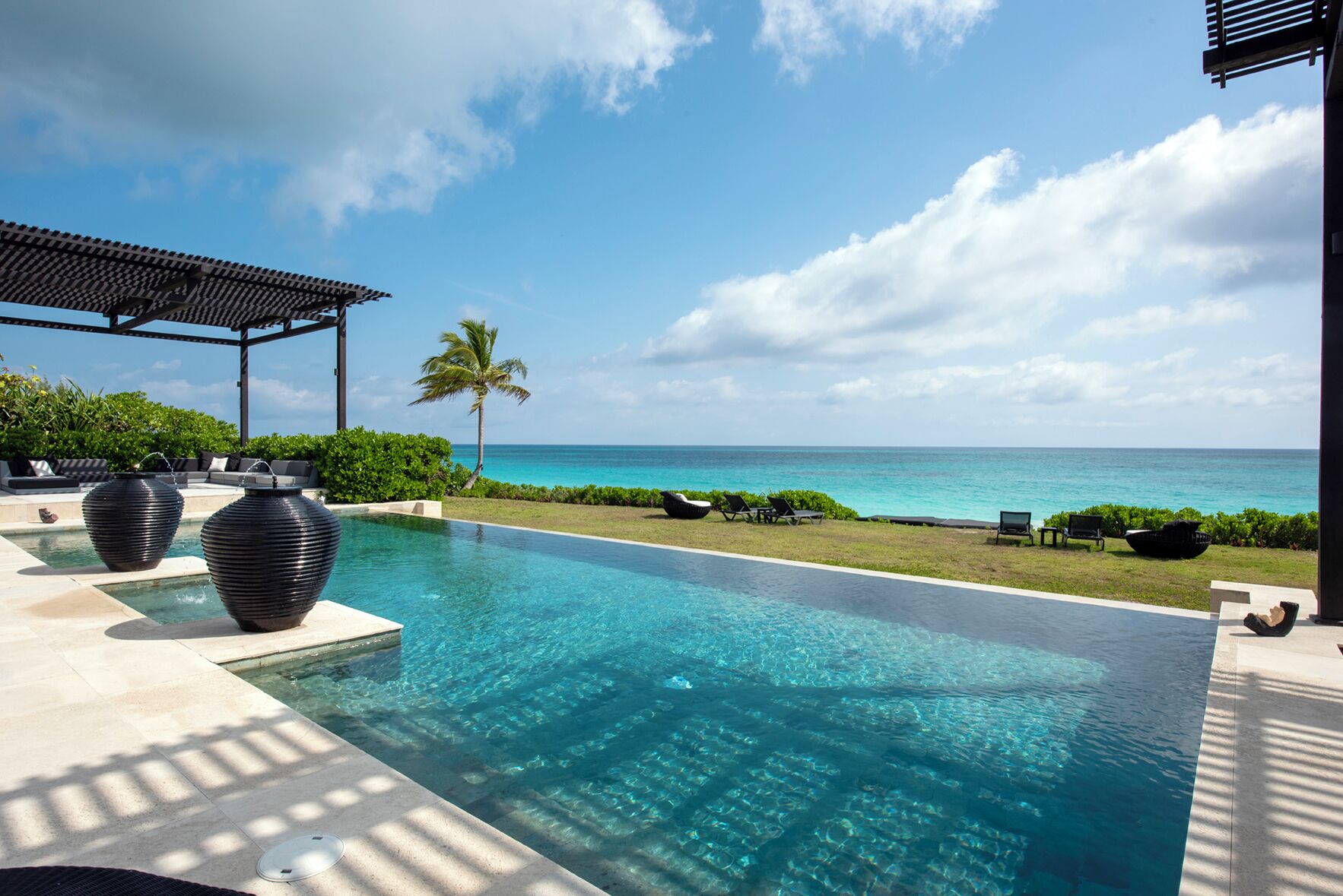 A clear blue pool with a view of the ocean, surrounded by lounge chairs and a palm tree.