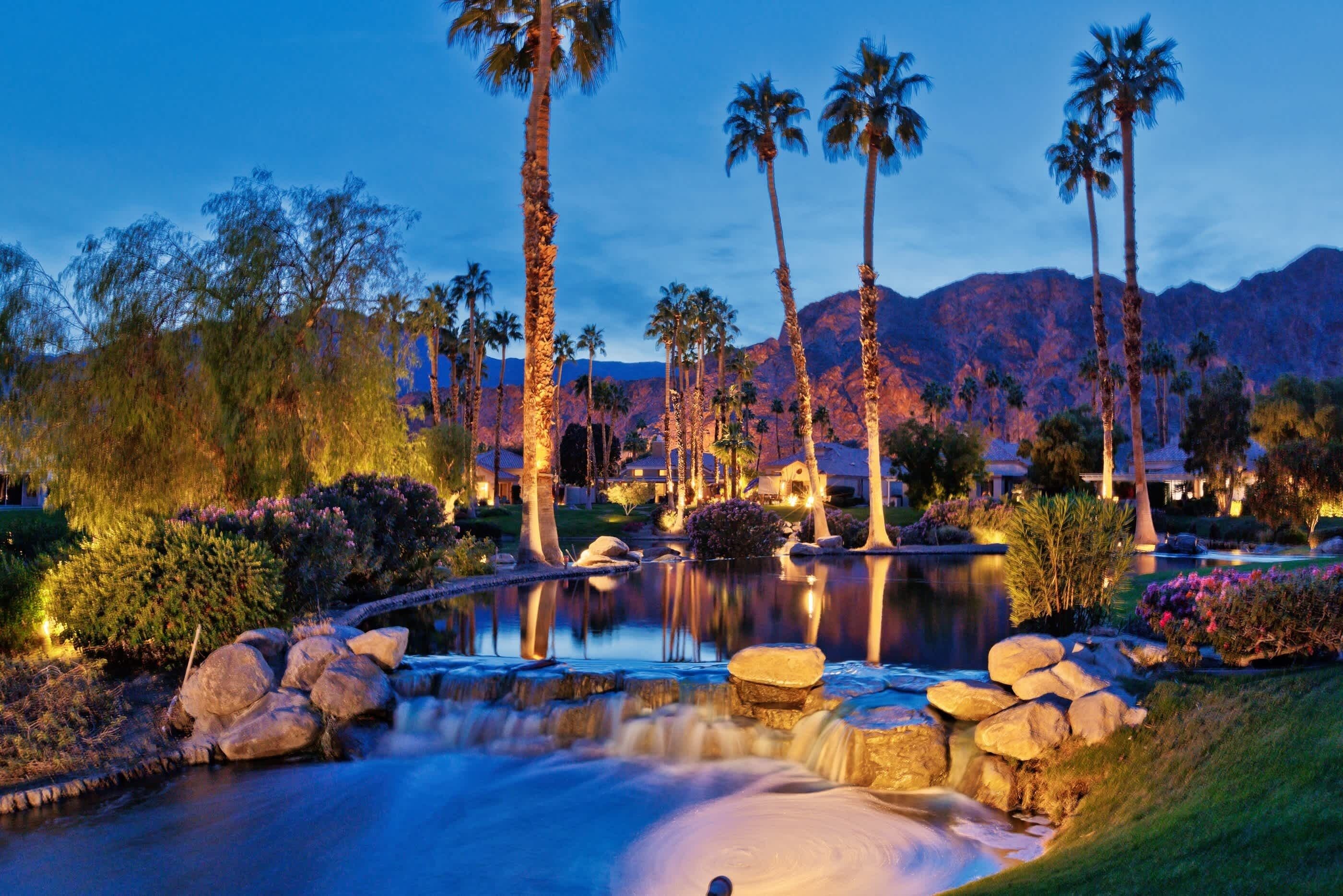 A serene landscape with a pond, palm trees, and mountains in the background.
