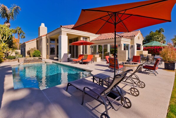 Un área de piscina con sillones de descanso y sombrillas, un patio con mobiliario exterior y una casa con tejado de baldosas.