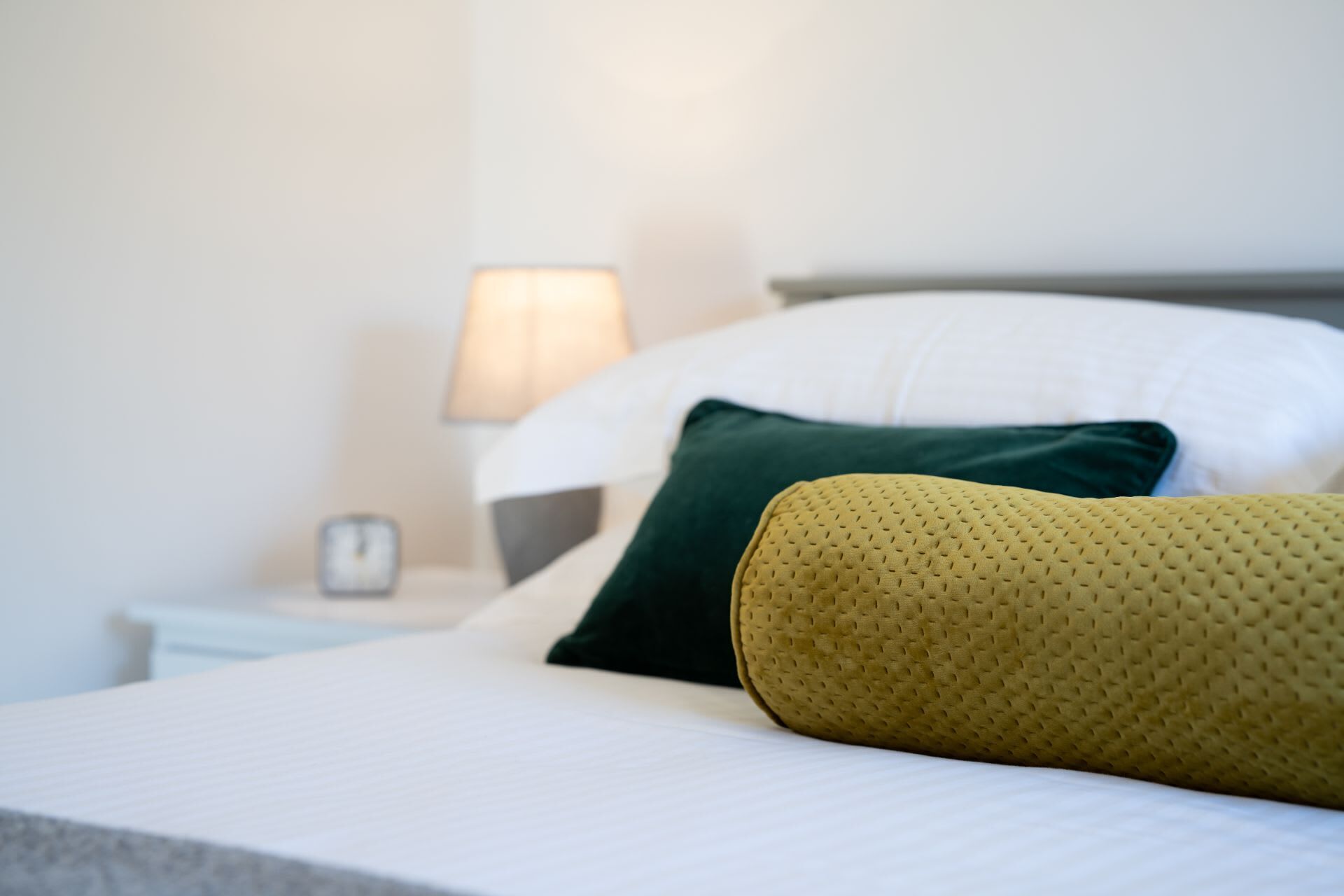 Cama bem arrumada com almofadas brancas e verdes, uma almofada de destaque amarela e uma mesa de cabeceira branca com luminária.