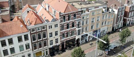 Una hilera de edificios de estilo europeo tradicional con tejados de tejas rojas, un cartel de hotel visible y una calle con peatones y bicicletas.