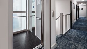 Um corredor moderno de hotel com elevador de vidro, piso coberto por carpete azul e corrimão branco.