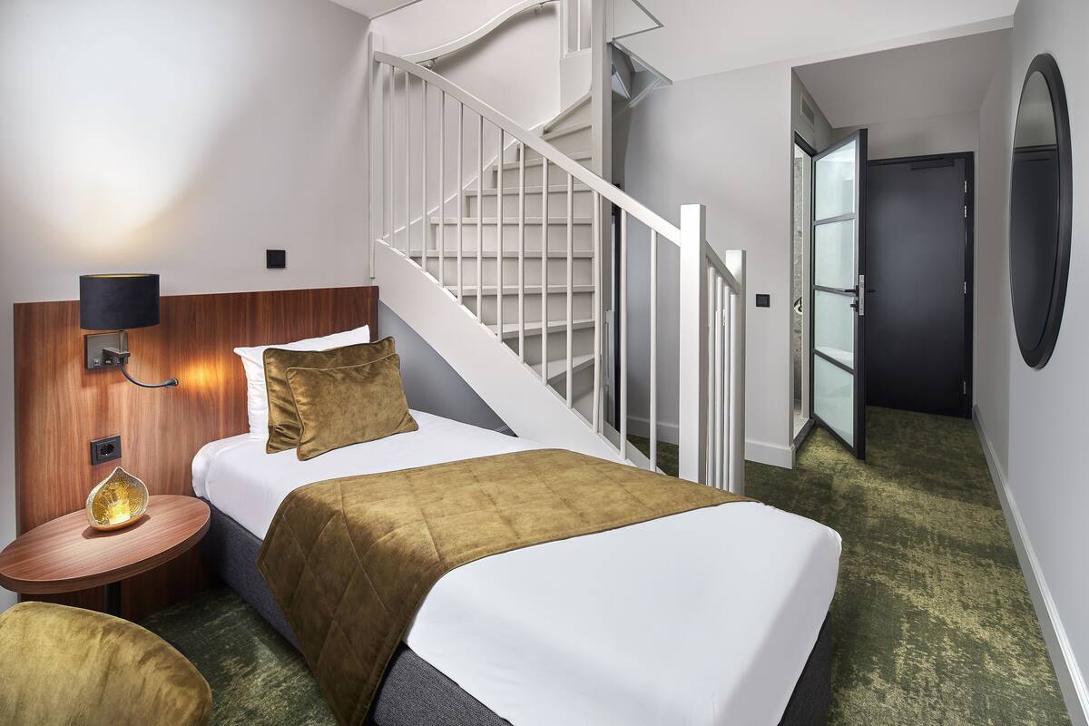 Una habitación de hotel con cama, mesita de noche, lámpara, escalera y puerta.