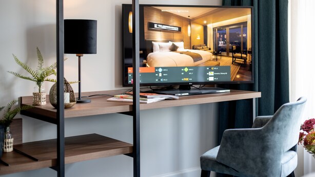 Quarto de hotel moderno com uma grande televisão de tela plana sobre um console de madeira, uma escrivaninha com luminária e uma cadeira confortável.