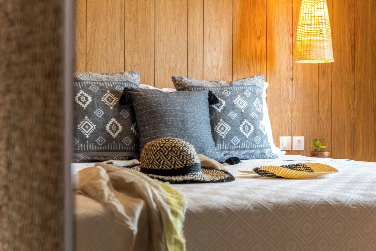 A neatly made bed with patterned pillows, a woven hat, and a small plant on a wooden wall.