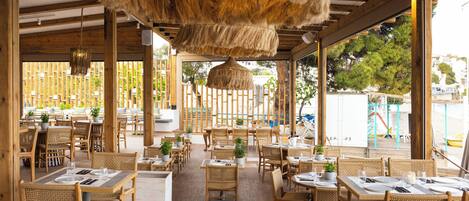 An outdoor dining area with wooden tables and chairs, potted plants, and a thatched ceiling.