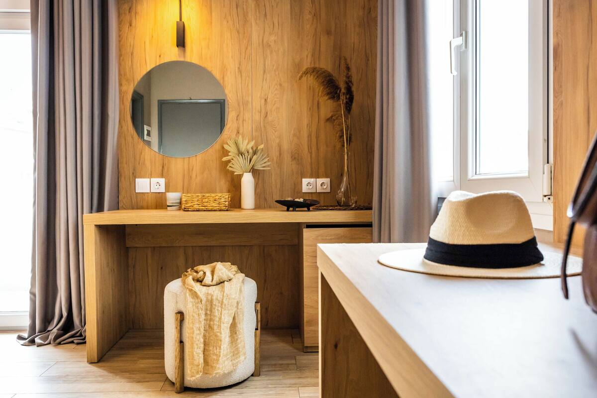 A wooden vanity with a round mirror, a vase with dried plants, and a hat on a table.