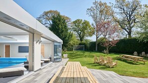 A modern outdoor patio with a pool, wooden tables, and chairs, surrounded by a well-maintained lawn and trees. - Finest House In Cheltenham ! (Cheltenham)