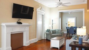 A living room with a fireplace, a flat-screen TV mounted above it, a ceiling fan, a sofa, a coffee table, and a dining area in the background.