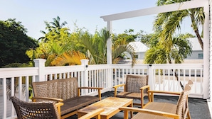 A covered outdoor seating area with wicker furniture and a wooden table, surrounded by lush tropical plants and a white fence.
