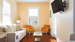 A living room with a sofa, two armchairs, a coffee table, a TV mounted on the wall, and a window with blinds.