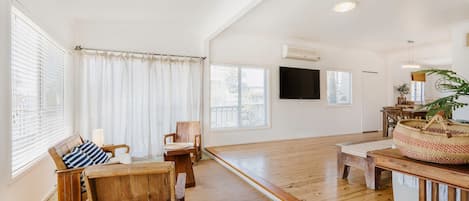 A spacious living room with wooden flooring, a sofa, a chair, a dining table, and a TV mounted on the wall.