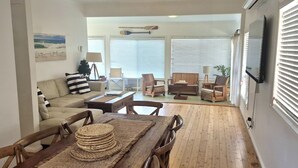 A living room with a sofa, chairs, a dining table with a woven placemat, and a television mounted on the wall. - Argyle Beach House by Book Jervis Bay (Vincentia)