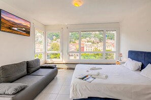 Habitación de hotel moderna con una cama grande, un sofá gris y vistas a un paisaje urbano a través de amplios ventanales.