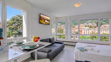 A modern hotel room with a large window, a sofa, a dining area with a table and chairs, and a bed with a view of the city.