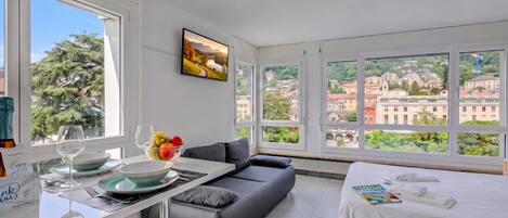 Habitación de hotel moderna con un ventanal grande, un sofá, un área de comedor con mesa y sillas, y una cama con vistas a la ciudad.