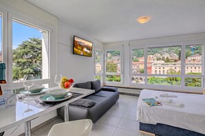 Habitación de hotel moderna con un ventanal grande, un sofá, un área de comedor con mesa y sillas, y una cama con vistas a la ciudad.