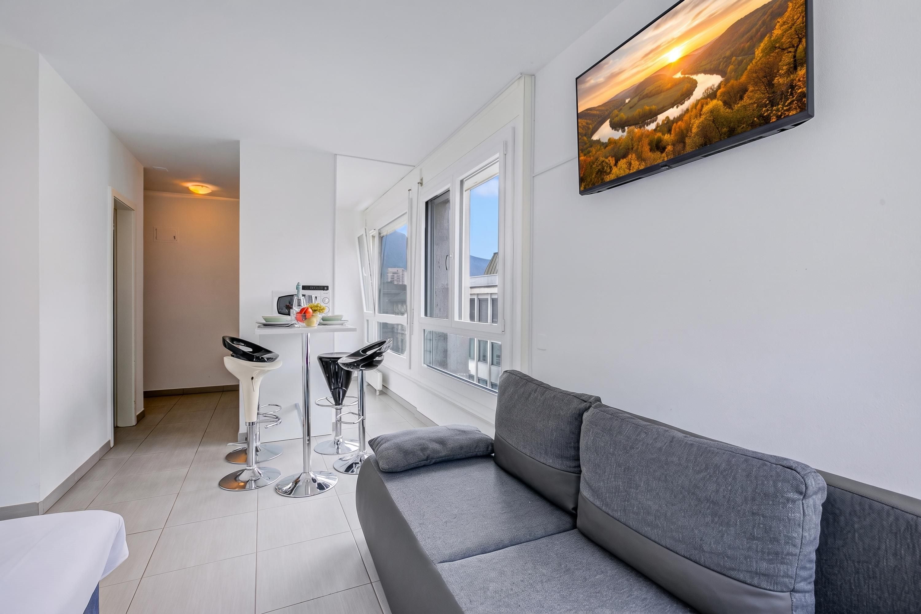 A modern living room with a grey sofa, two bar stools, a small table with a vase, and a large window.