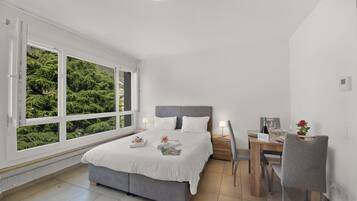 A modern bedroom with a large window, a bed, bedside tables, a desk, and a chair.