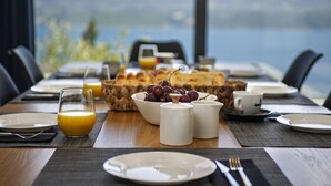 Una mesa de desayuno servida con platos, un vaso de jugo de naranja, un bol de uvas y una jarra de leche, con vistas a un paisaje pintoresco.