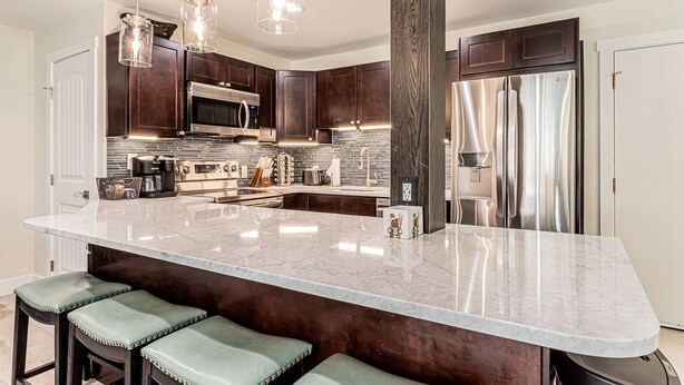 A modern kitchen with dark wood cabinets, stainless steel appliances, a marble island with bar stools, and pendant lighting. - Remodeled 1-Bdrm Ski-In/-Out Condo w/ Fireplace! (Steamboat Springs)
