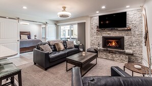 A living room with a stone fireplace, a black leather sofa, a coffee table, and a flat-screen TV mounted on the wall. - Remodeled 1-Bdrm Ski-In/-Out Condo w/ Fireplace! (Steamboat Springs)