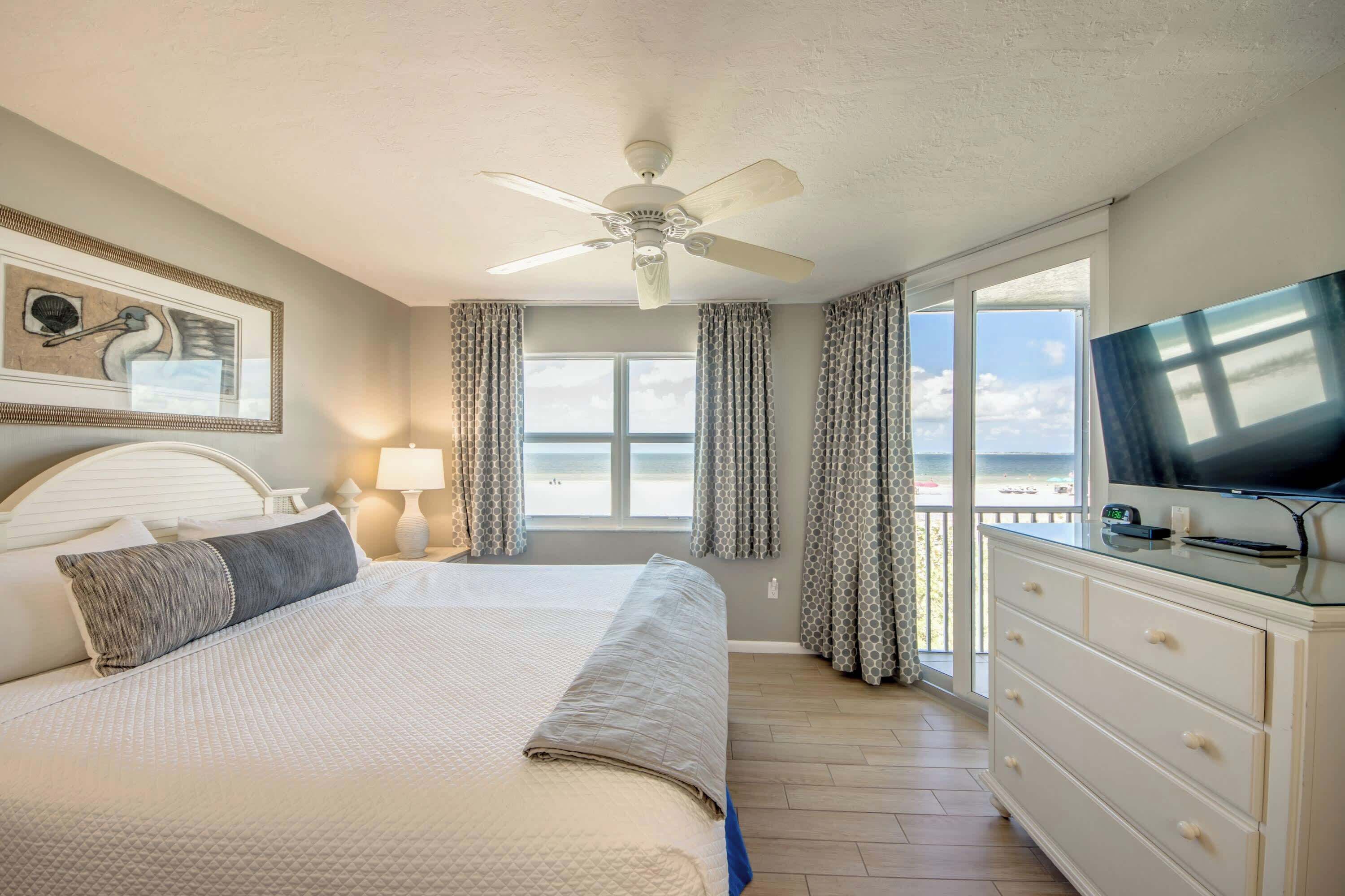 A hotel room with a bed, a television, a desk, and a view of the beach.