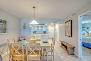 A dining area with a wooden table and chairs, a hanging light fixture, and a kitchen in the background.