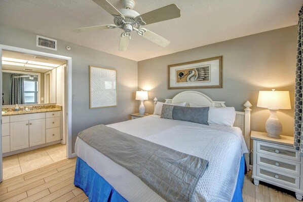A bedroom with a bed, two bedside tables with lamps, a ceiling fan, a large mirror, and artwork on the wall.