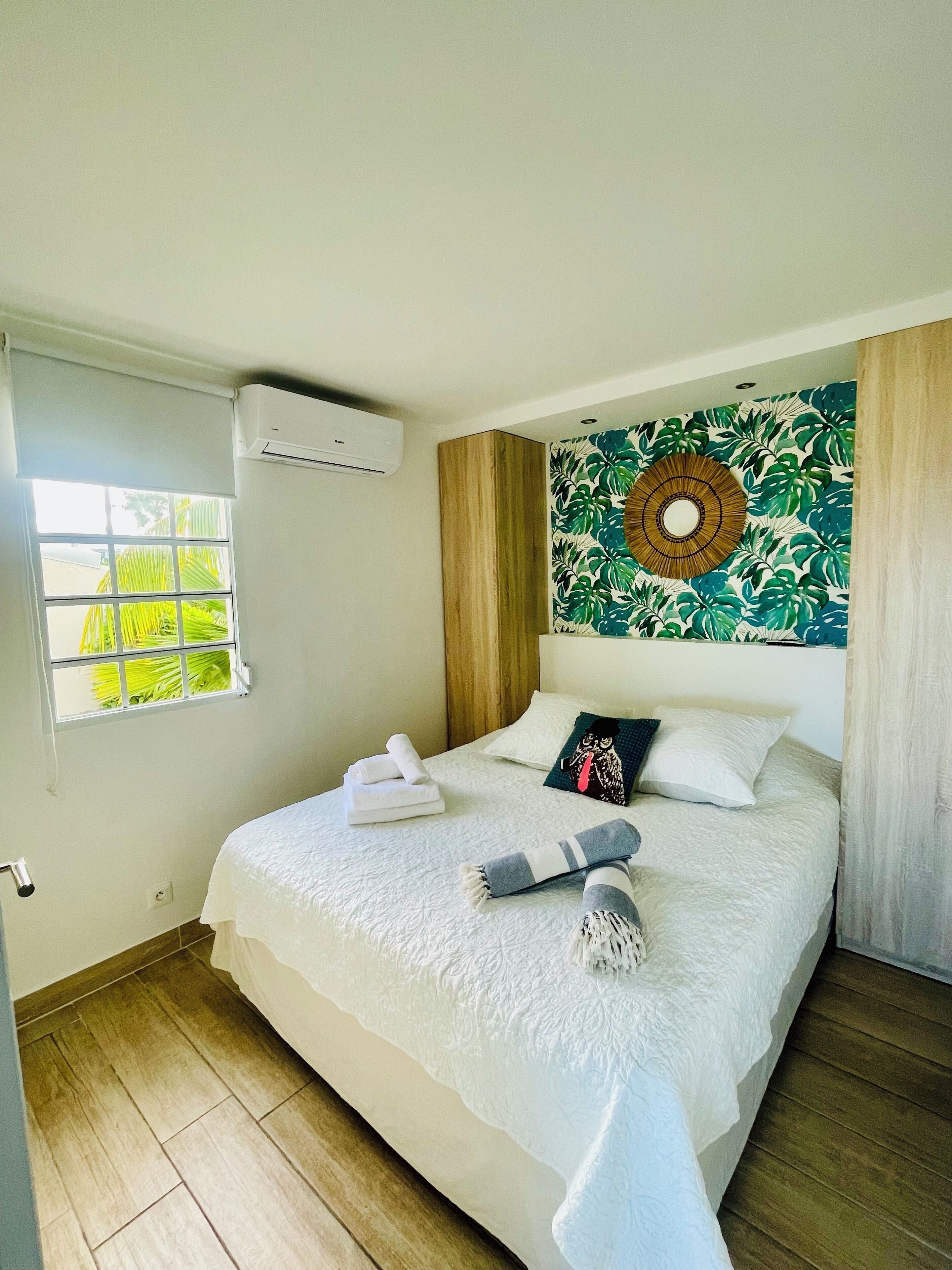 A neatly made bed with towels, a patterned wall hanging, and a window with blinds.