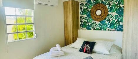 A neatly made bed with towels, a patterned wall hanging, and a window with blinds.