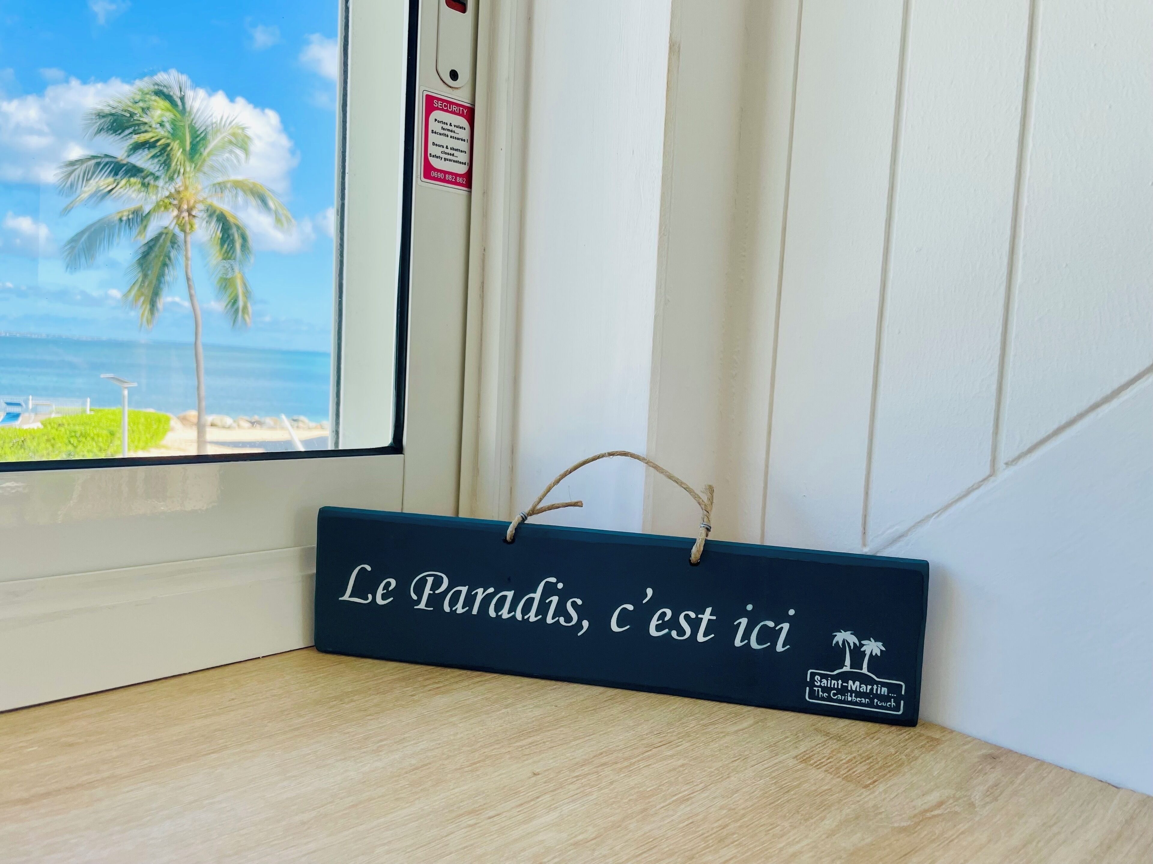 Une enseigne en bois portant l’inscription « Le paradis, c’est ici » et ornée d’un logo représentant un palmier, placée sur un plancher en bois près d’une fenêtre donnant sur une plage.