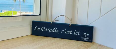 Une enseigne en bois portant l’inscription « Le paradis, c’est ici » et ornée d’un logo représentant un palmier, placée sur un plancher en bois près d’une fenêtre donnant sur une plage.