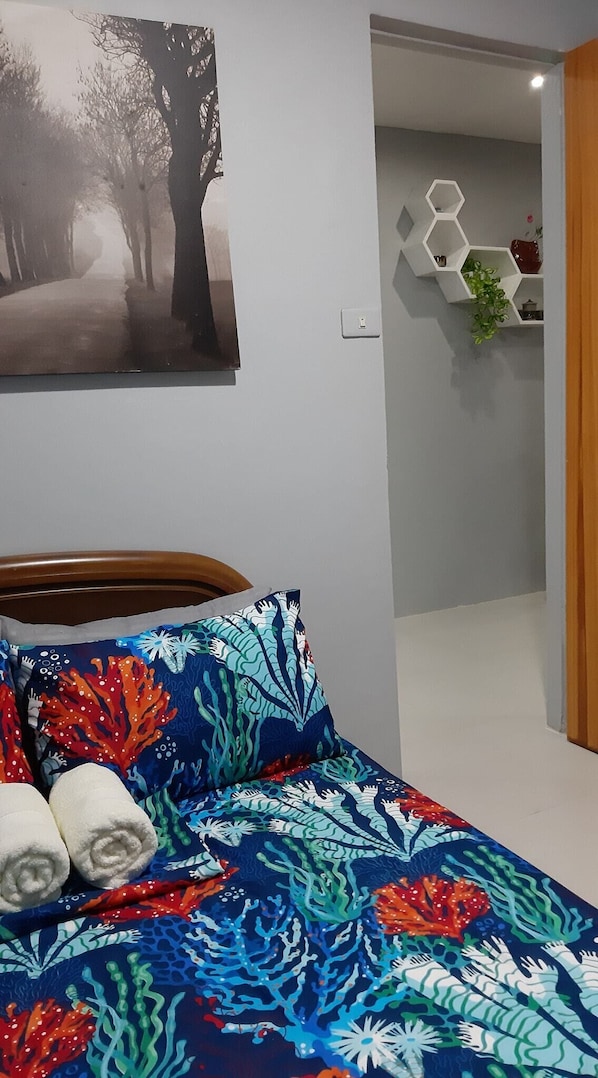 A bedroom with a bed featuring a colorful coral pattern, a painting of trees, and a hexagonal shelf with plants.